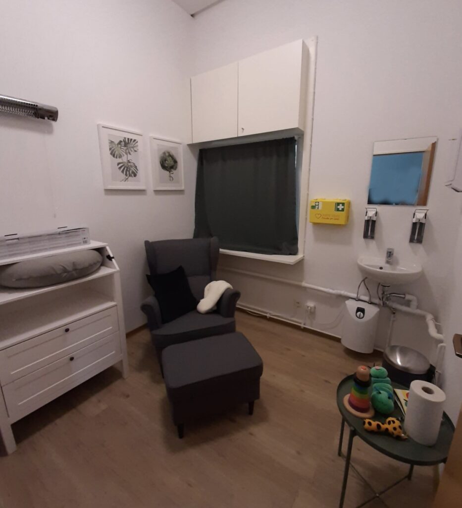 Ansicht des Stillraums mit Wickeltisch am linken Bildrand. Über dem Wickeltisch ist eine Wärmelampe angebracht. Neben dem Wickeltisch steht ein Ohrensessel mit Kissen und Decke, davor ein Hocker. Das hinter dem Sessel befindliche Fenster ist mit einem Vorhang abgedunkelt. Rechts vom Sessel ist ein Waschbecken mit Spiegel angebracht Am rechten Bildrand befindet sich ein kleiner Beistelltisch, auf dem Spielzeug, ein Buch und Papiertücher ausgelegt sind.