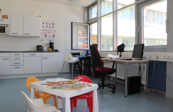 Raumansicht des Eltern-Kind Arbeitsraums mit Kindertisch im Vordergrund. Im Hintergrund steht ein Arbeitstisch mit einem Bürostuhl vor einer Fensterfront. Der Arbeitsplatz ist mit PC, Bildschirm, Tastatur und Maus ausgestattet. Vor dem Schreibtisch ist eine Steckdosenleiste angebracht. Neben dem Schreibtisch befindet sich ein kleiner Esstisch mit Klappstühlen und einem Hochstuhl. Die Küche mit Wasserkocher, Kaffeemaschine, Mikrowelle und diversen Schränken ist auf der rechten Seite im Hintergrund abgebildet.