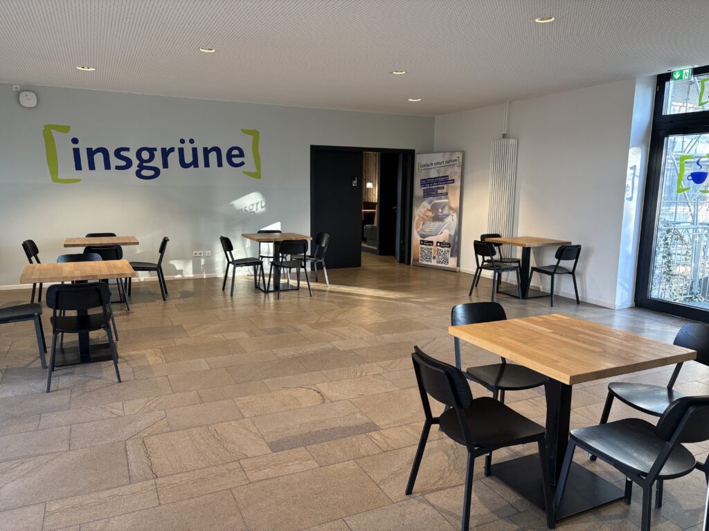 Aufenthaltsraum hinter dem Verkaufsraum mit kleinen viereckigen Tischen, die jeweils mit vier Stühlen bestuhlt.