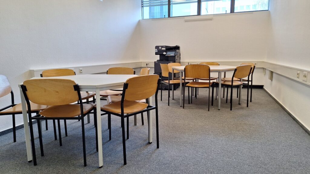 Arbeitsraum mit zwei sechseckigen Tischen mit jeweils sechs Stühlen. An den Wänden sind Steckdosenleisten angebracht. Kopiergerät im Hintergrund sowie Fenster im oberen Bildrand.
