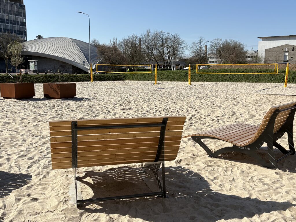 Zwei Sonnenliegen, die am Rand zweier Beachvolleyballfelder auf Sandboden aufgestellt sind. Im Hintergrund sind zwei aufgestellte Beachvolleyball-Netze, Blumenbeete sowie angrenzende Gebäude zu sehen.