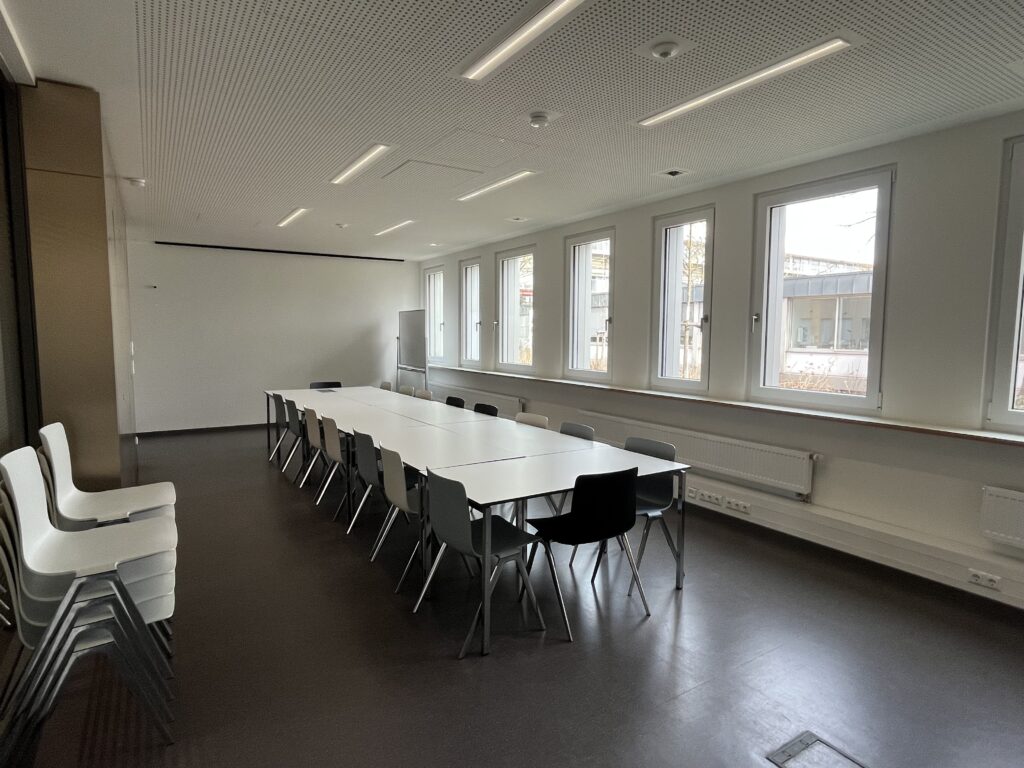 Raumansicht vom Eingangsbereich aus fotografiert. Eine weiße, längliche Tischgruppe im Vordergrund, umgeben von Stühlen. Auf der linken Seite sind fünf Fenster zu sehen. Ein Stapel zusätzlicher Stühle steht links im Vordergrund. Im Hintergrund ist eine große weiße Projektionsfläche. An den Fußleisten befinden sich Steckdosen.