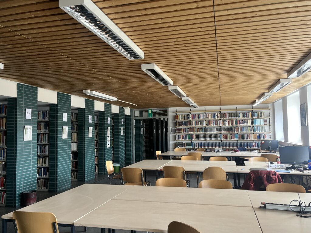 Bibliotheksraum mit zusammengestellten Tischen mit Stühlen. Die Tische sind mit Mehrfachstechdosen ausgestattet. An jedem der drei Tische befindet sich neben ca. 8 einfachen Arbeitsplätzen auch ein Arbeitsplatz mit PC, Monitor, Tastatur und Maus. Bücherregale im Bildhintergrund sowie auf dem linken Bildrand.